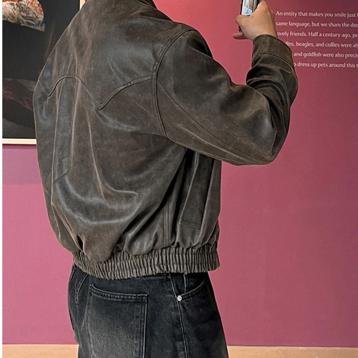 Person wearing a brown leather jacket taking a photo with a smartphone against a pink wall.
