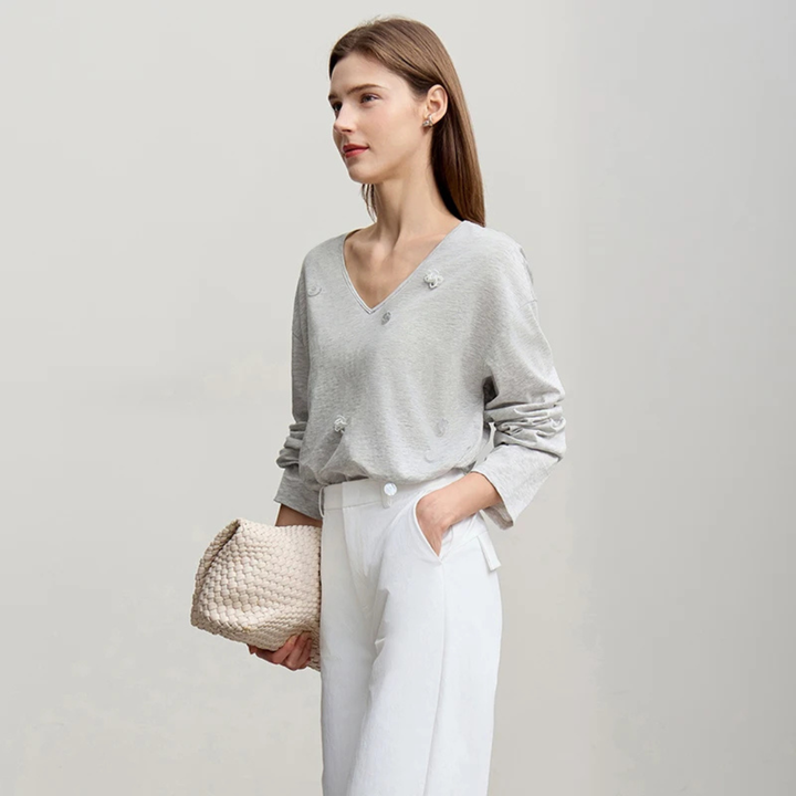 Woman wearing a gray sweater and white pants holding a beige handbag on a light gray background