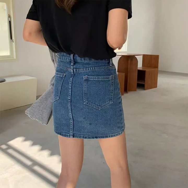 Person wearing a black top and blue denim skirt in a minimalistic room.
