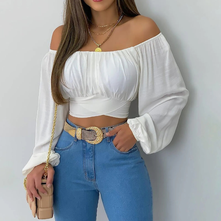 Woman wearing a white off-shoulder top and blue jeans with a gold belt, holding a beige handbag.