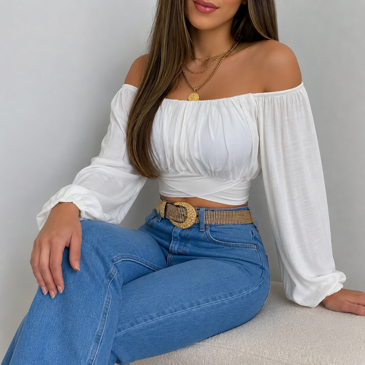 Woman wearing a white off-shoulder top and blue jeans sitting against a plain background