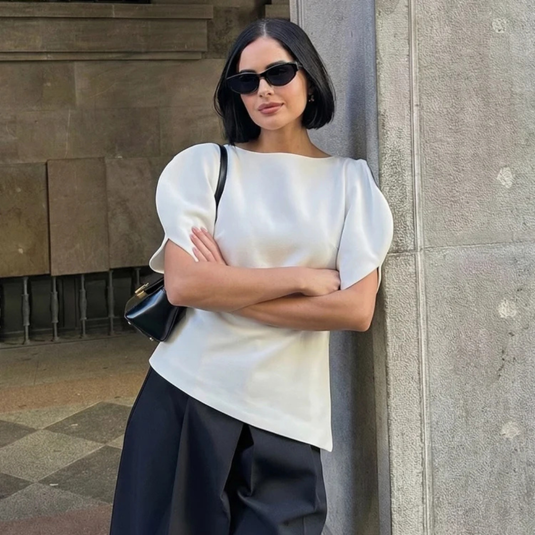 Woman in a white top and black pants standing against a wall.