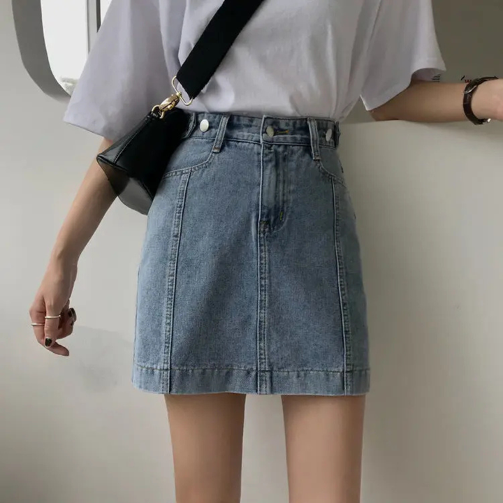 Person wearing a white shirt and blue denim skirt with a black bag on a plain background
