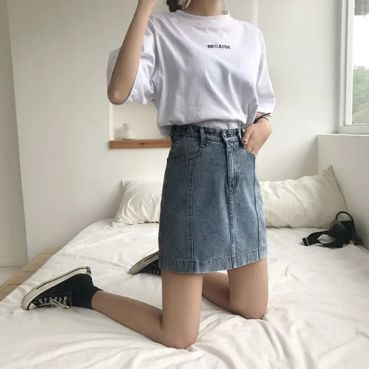 Person wearing a white t-shirt and blue denim skirt sitting on a bed.