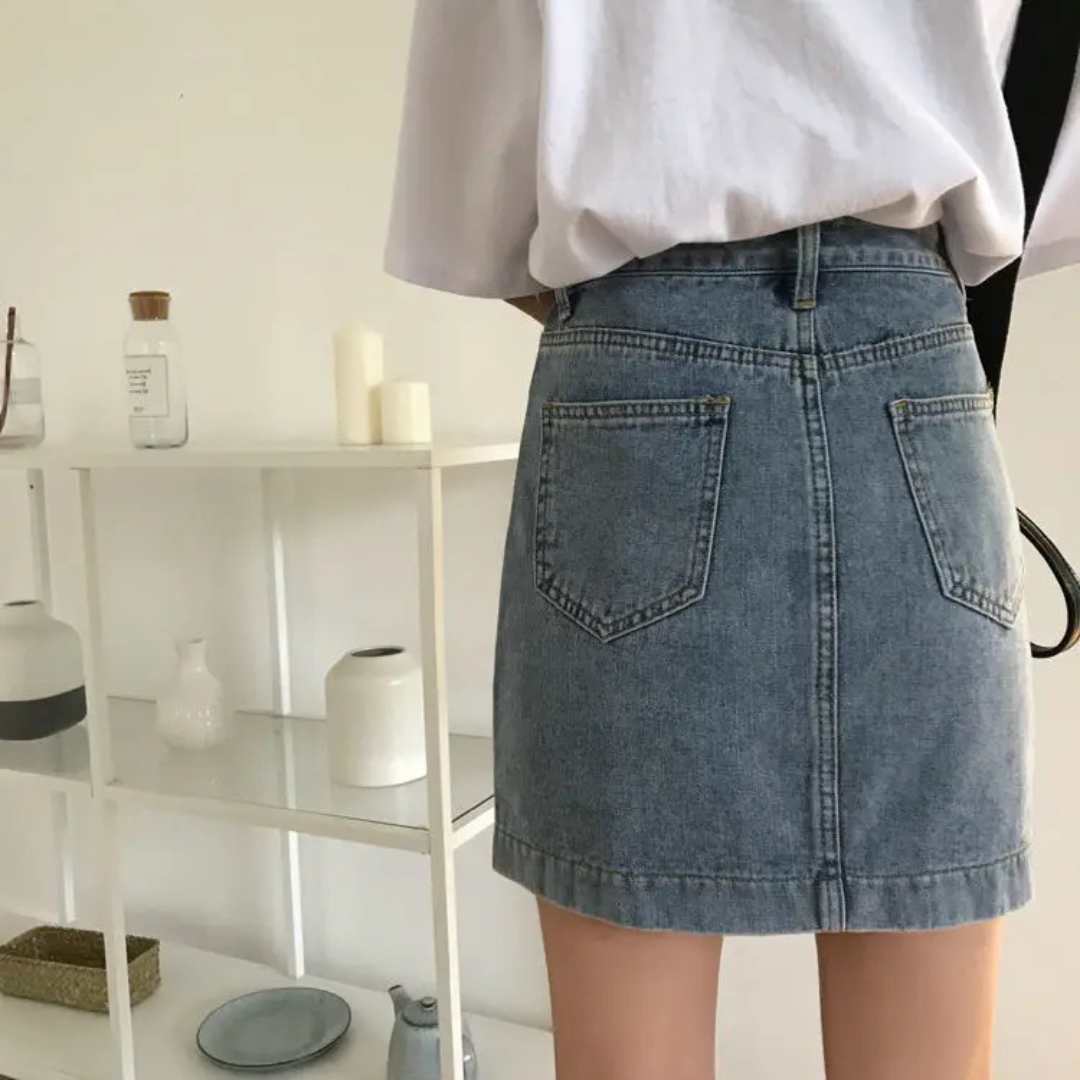 Person wearing a blue denim skirt with a white shirt in a minimalistic room.