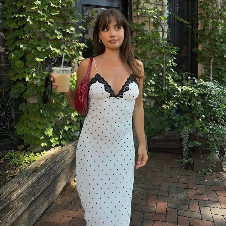 Woman in a white polka dot dress with black lace details, holding a drink, standing outdoors.