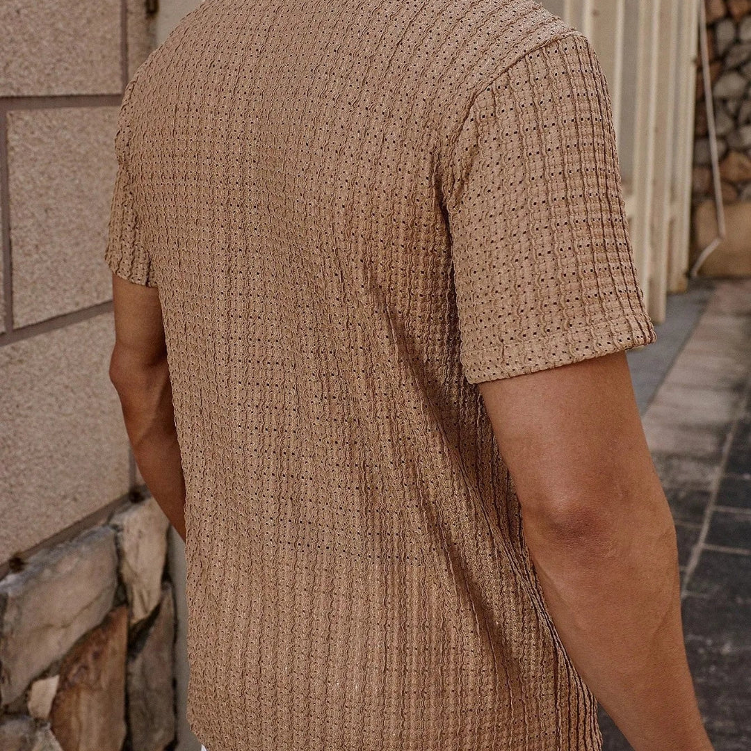 Person wearing a khaki textured shirt standing against a stone wall.