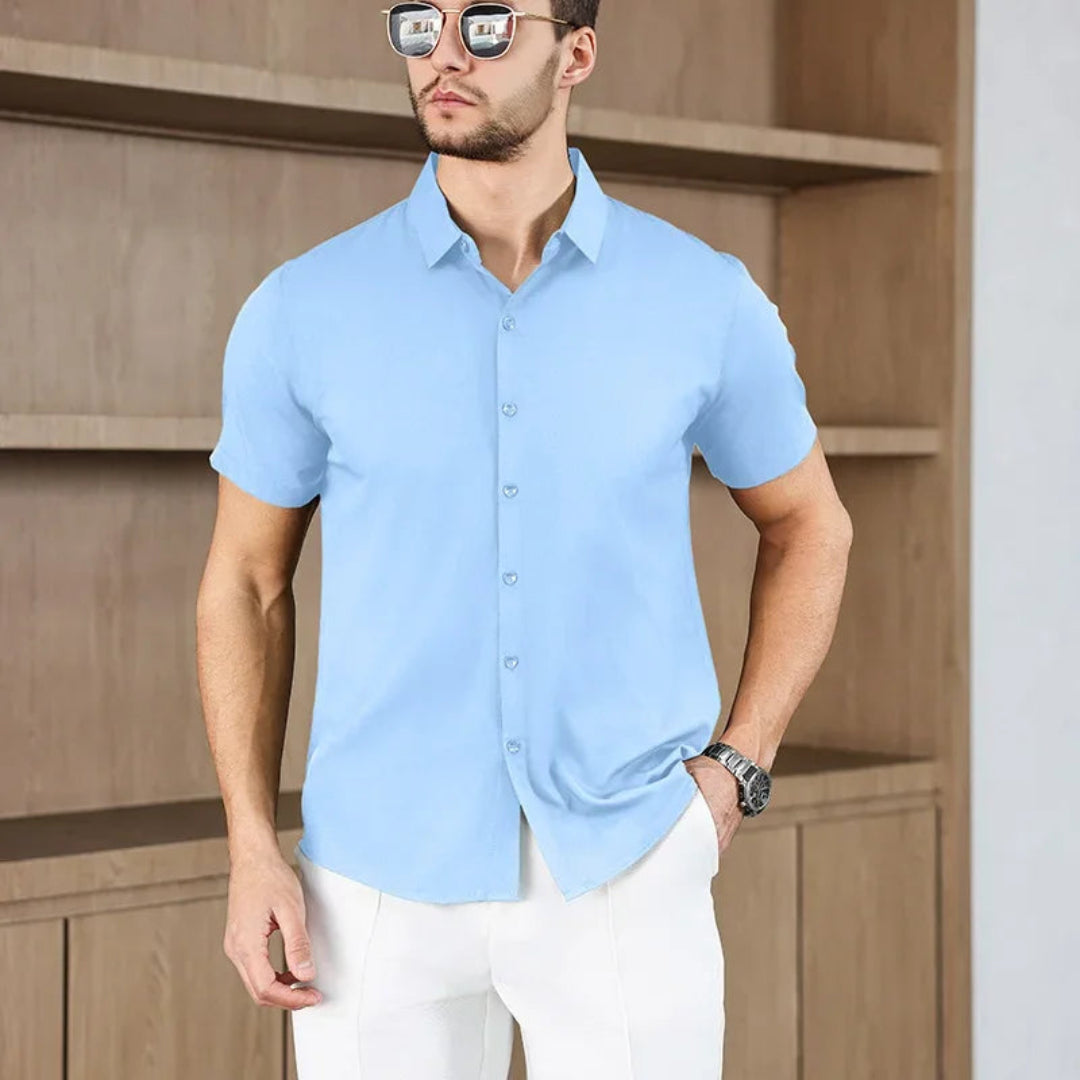 Man wearing a blue short-sleeve shirt and white pants standing against a wooden background.