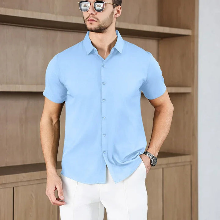 Man wearing a blue short-sleeve shirt and white pants standing against a wooden background.