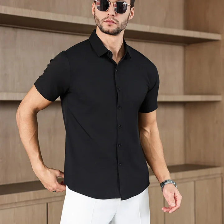 Man wearing a black short-sleeve shirt and sunglasses against a beige wall.