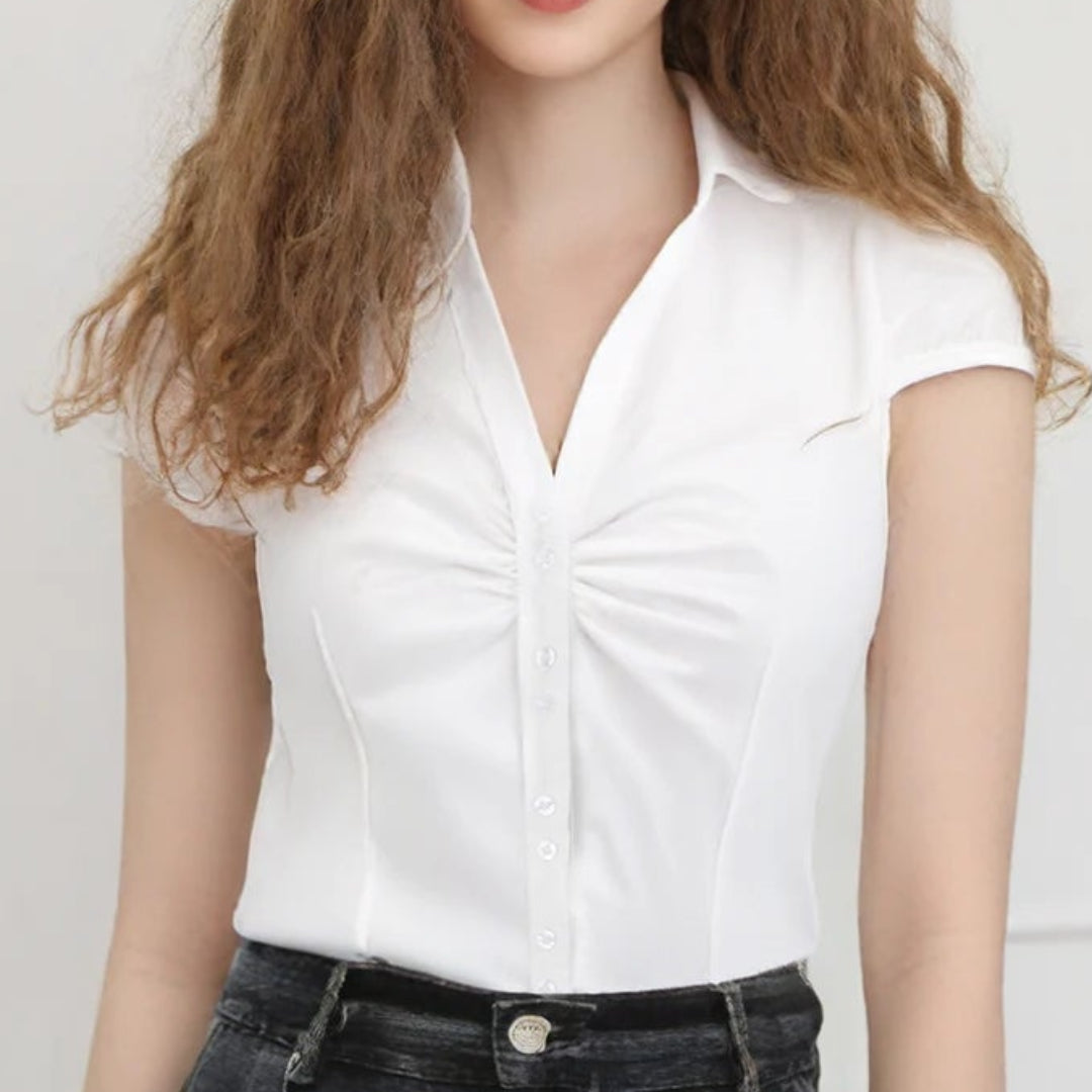 Woman wearing a white blouse and black shorts standing against a white wall with a vase of flowers on a table.