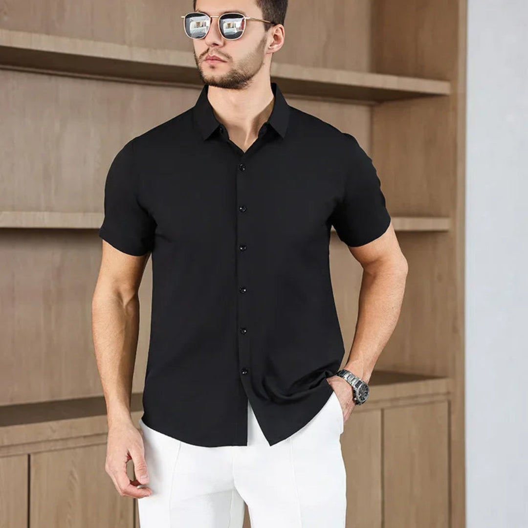 Man wearing a black short-sleeve shirt and white pants against a wooden wall.