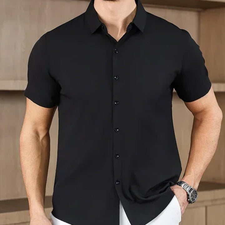 Man wearing a black short-sleeve shirt and white pants against a wooden wall.