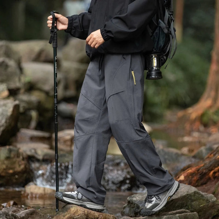 Person hiking in a forest wearing black jacket and gray pants with a backpack and walking stick.