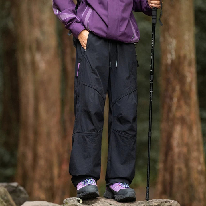 Person wearing a purple jacket and black pants with hiking boots, standing on a rock in a forest.