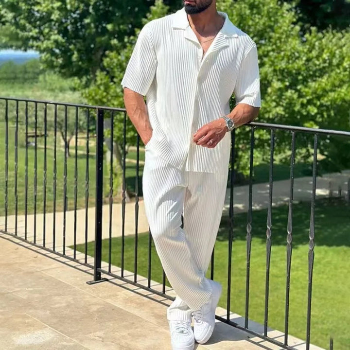 Man in a white outfit standing on a balcony with greenery in the background
