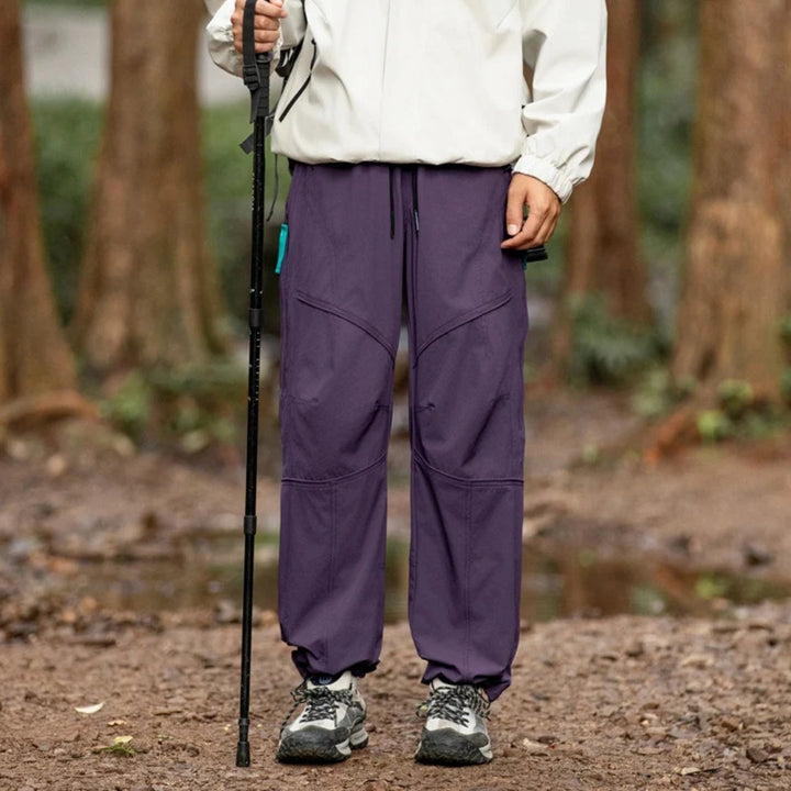 Person wearing purple hiking pants and a white jacket with a walking stick in a forest setting.