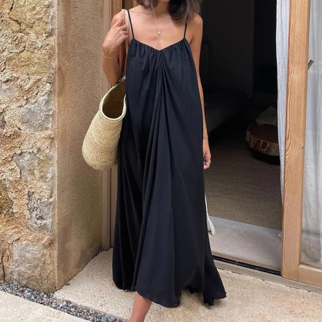 Woman in a Selene Backless Maxi Dress holding a straw bag against a stone wall.
