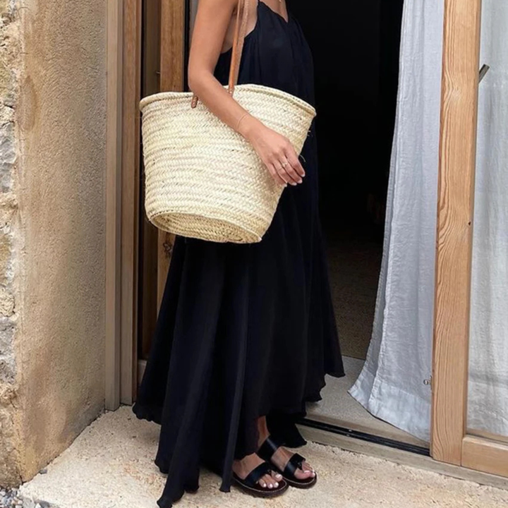 Person wearing a Selene Backless Maxi Dress holding a straw bag, standing in front of a door.