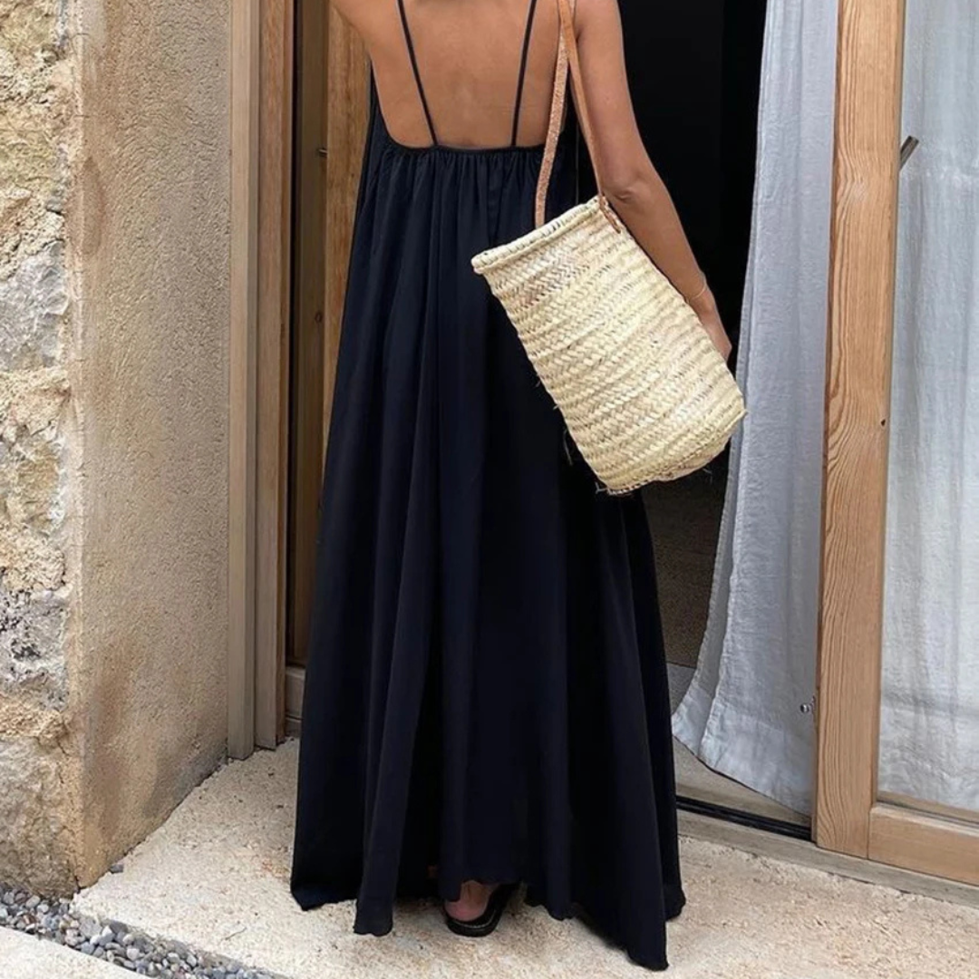 Woman in a Selene Backless Maxi Dress holding a straw bag, standing in front of a stone wall.