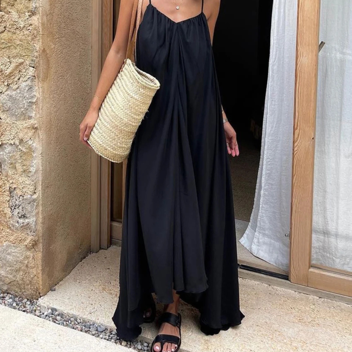 Woman in a Selene Backless Maxi Dress holding a straw bag, standing in front of a stone wall.