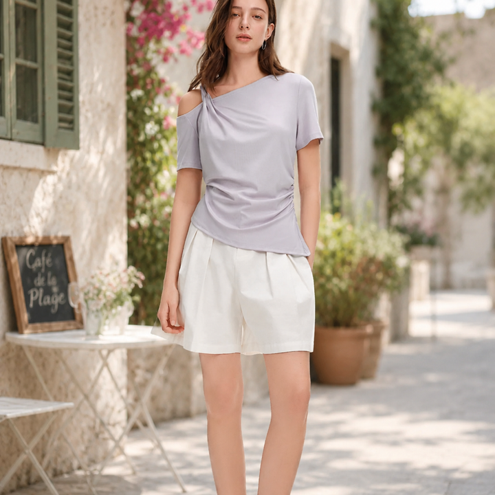 Woman in a light purple off-shoulder top and white shorts standing in a sunlit outdoor setting.
